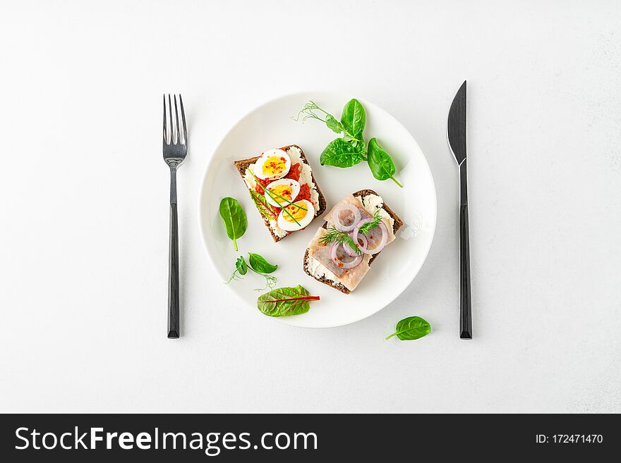 Danish smorrebrod open sandwiches on white plate, fork and knife, green salad leaves on white background