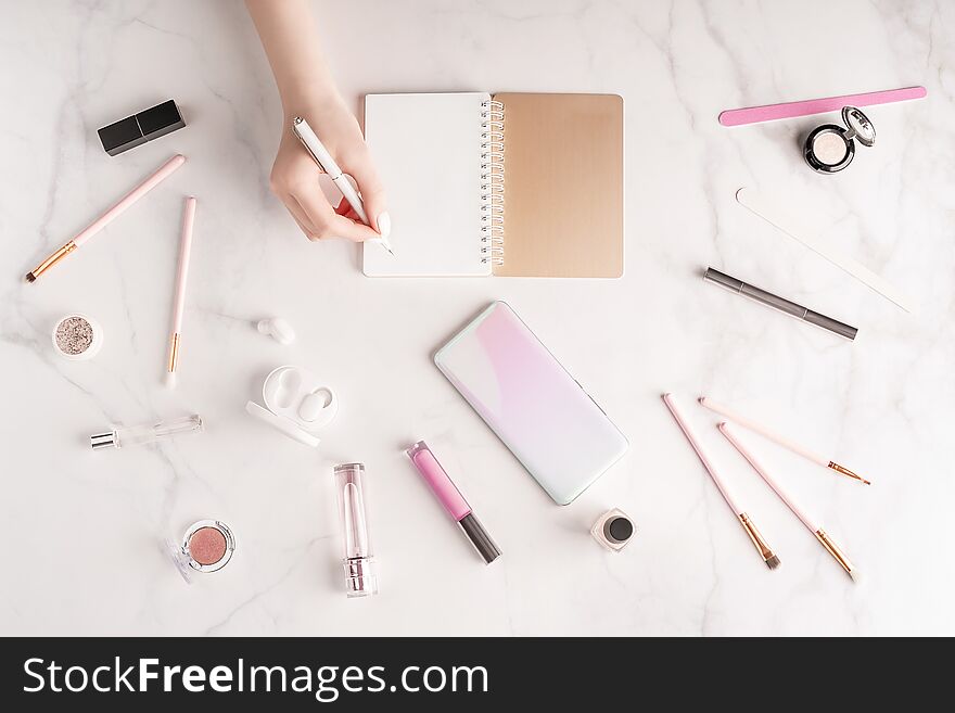 Feminine workspace in flat lay style with female hand writing in notepad, up-to-date smartphone mockup, cosmetics and