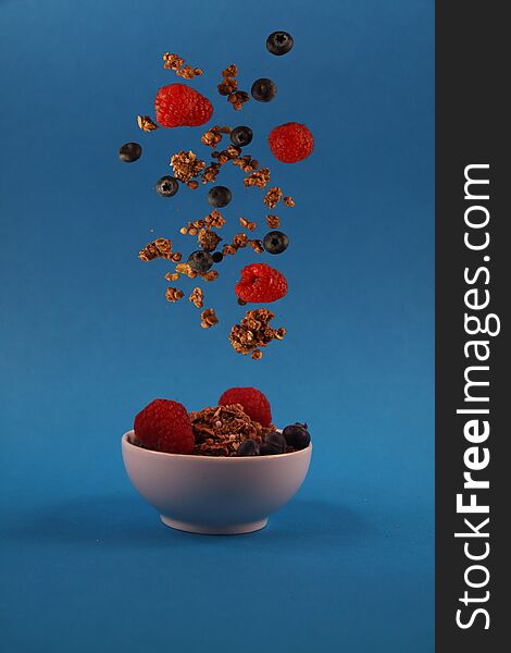 Granola and berries pouring to the white bowl against blue background. Image contains copy space. Concept of flying food