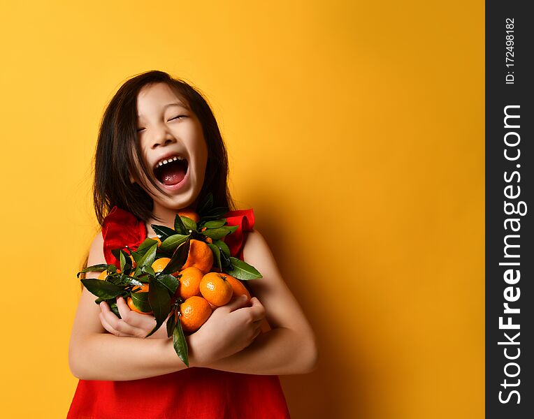 Little asian child in a red blouse. Laughing out loud with a toothless mouth, holding tangerines and oranges in their hands with green leaves, posing on an orange background. Childhood, emotions. Little asian child in a red blouse. Laughing out loud with a toothless mouth, holding tangerines and oranges in their hands with green leaves, posing on an orange background. Childhood, emotions.