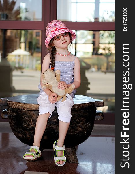 Small girl in glasess and sun hat on vacation sits with her toy