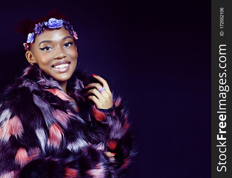 Young pretty african american woman in spotted fur coat and flowers on head smiling on black background, fashion people