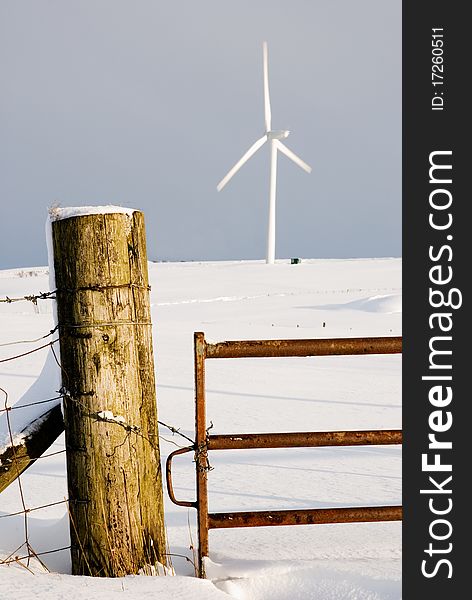 A vertical image of a winter snowscene with gate to a field leading to a wind turbine. A vertical image of a winter snowscene with gate to a field leading to a wind turbine