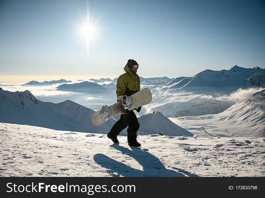 Guy with snowboard in winter clothes walks on high snowy sunny mountain top at sunny day. Guy with snowboard in winter clothes walks on high snowy sunny mountain top at sunny day