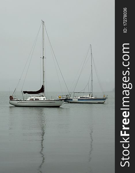 Two boats sit on a lake surrounded by mist and the reflections of their masts on the water. Two boats sit on a lake surrounded by mist and the reflections of their masts on the water