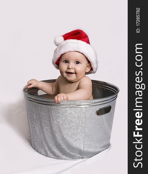 Smiling baby wearing a Santa hat, holds on to the sides of a wash basin while sitting in it. Smiling baby wearing a Santa hat, holds on to the sides of a wash basin while sitting in it.