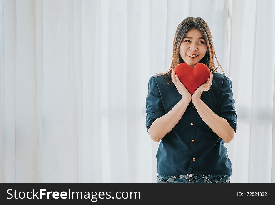 Smiling Asian woman holding and showing red heart in white room. Love and valentines day concept. Smiling Asian woman holding and showing red heart in white room. Love and valentines day concept