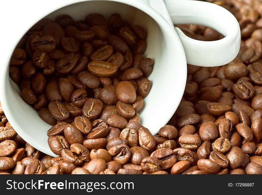 A white cup surrounded and partly filled with coffee beans. A white cup surrounded and partly filled with coffee beans