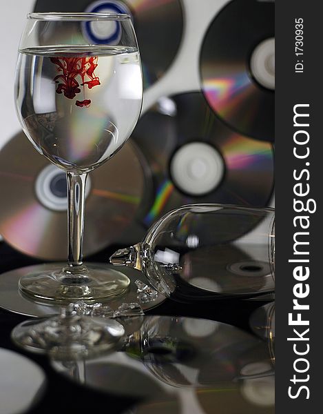 A shot of a broken wine glass and another with drops of red inside and a background of compact disks. A shot of a broken wine glass and another with drops of red inside and a background of compact disks.