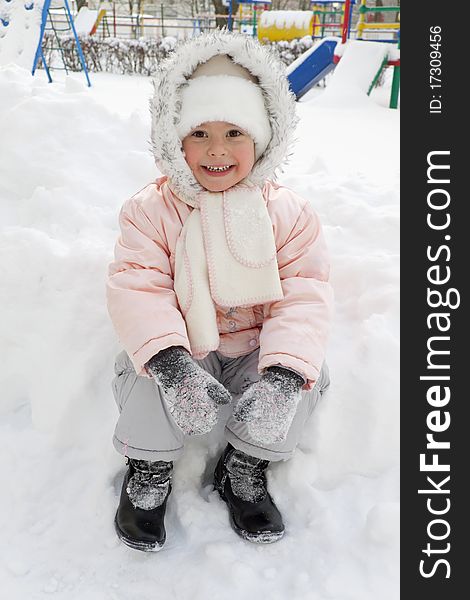 Warmly dressed joyful child sits in a snowdrift on a playground. Warmly dressed joyful child sits in a snowdrift on a playground