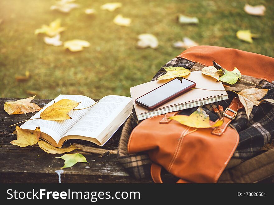 The backpack is placed on an antique wooden table with books, notebooks, phones and the nature of maple leaves from the top view