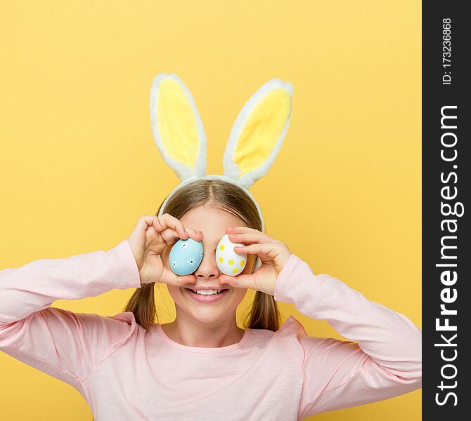 Cheerful child with bunny ears covering eyes with easter eggs isolated on yellow