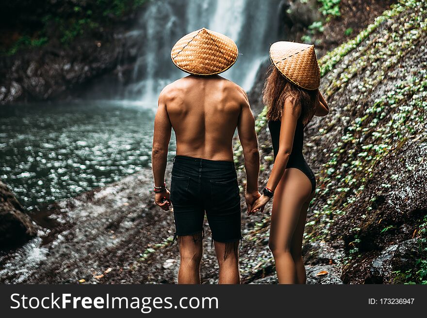 Lovers At The Waterfall, Rear View. Couple Admiring A Beautiful Waterfall In Indonesia. Couple On Vacation In Bali. Honeymoon Trip