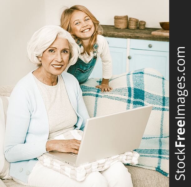Smiling Granny And Her Granddaughter Using Wireless Internet At Home Mature Woman With Blond Hair Sitting On Sofa And Typing On Computer, Lady Working Or Messaging With Someone On Laptop Computer. Smiling Granny And Her Granddaughter Using Wireless Internet At Home Mature Woman With Blond Hair Sitting On Sofa And Typing On Computer, Lady Working Or Messaging With Someone On Laptop Computer