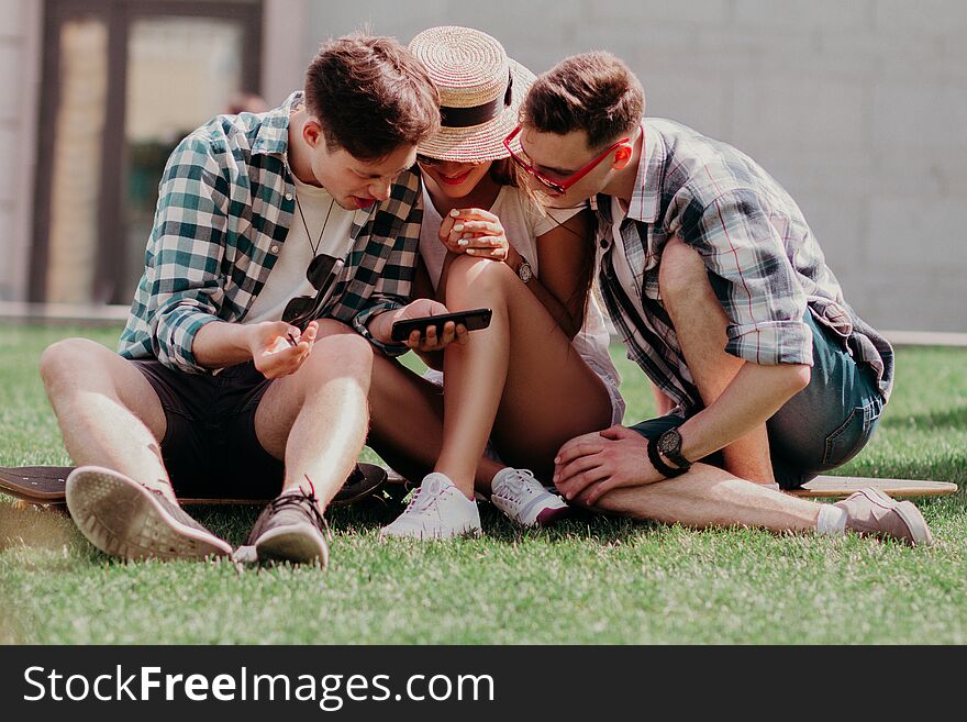 The Youngsters Leaned Together Over A Mobile Choosing The Best Photo Sitting On The Grass On A Summer Day. Stylish Young People In Casual. The Youngsters Leaned Together Over A Mobile Choosing The Best Photo Sitting On The Grass On A Summer Day. Stylish Young People In Casual.