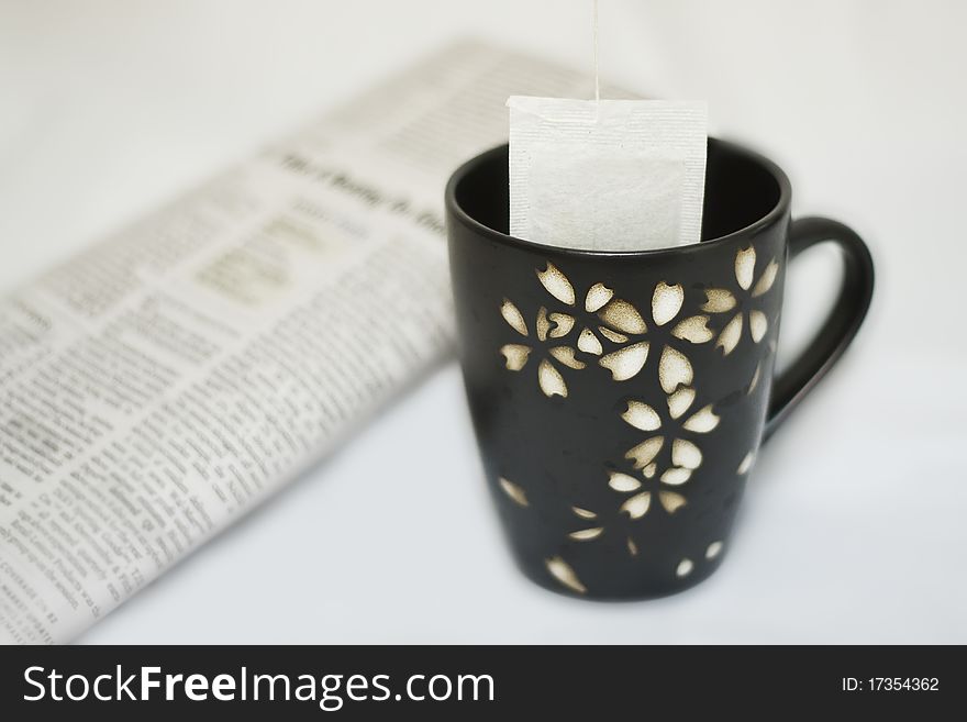 Tea cup with a tea bag hanging from above, and a newspaper in soft focus in background. Tea cup with a tea bag hanging from above, and a newspaper in soft focus in background