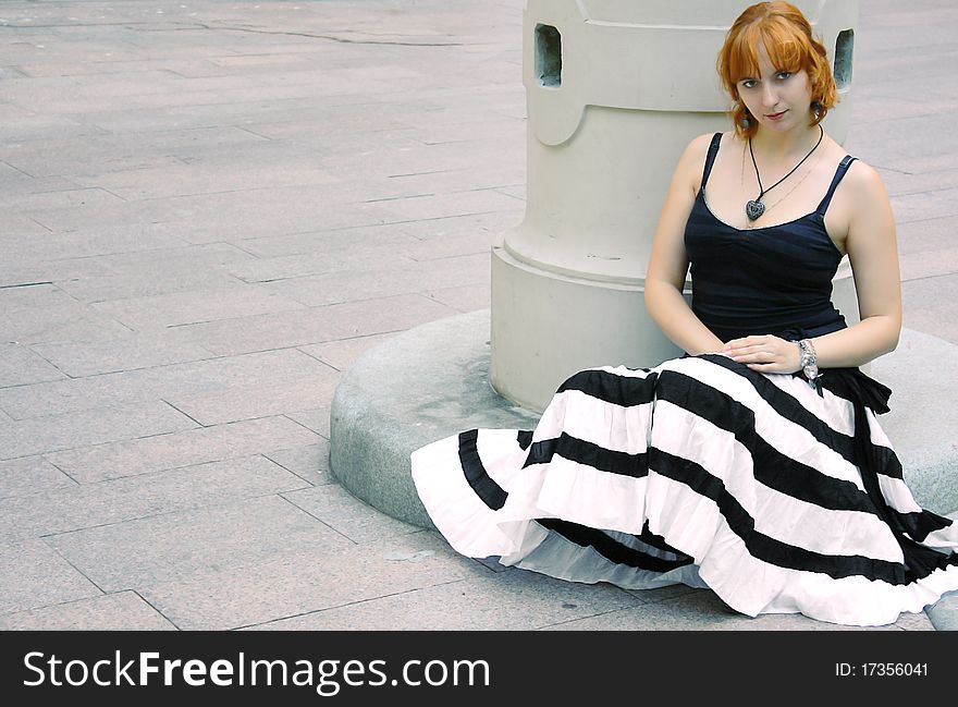 Young red woman is sitting in flared striped black and white skirt. She has vintage pendant and big bracelet. Young red woman is sitting in flared striped black and white skirt. She has vintage pendant and big bracelet.