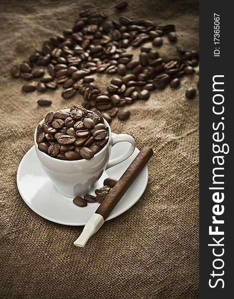 White ceramic cup with coffee beans and cigar on the background of burlap. White ceramic cup with coffee beans and cigar on the background of burlap