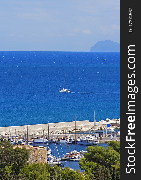 Small town marina, with a yacht leaving towards open sea, Mediterranean vegetation, ancient tower and seaside mountain in the distance Location: San Vito Lo Capo, Sicily, Italy. Small town marina, with a yacht leaving towards open sea, Mediterranean vegetation, ancient tower and seaside mountain in the distance Location: San Vito Lo Capo, Sicily, Italy