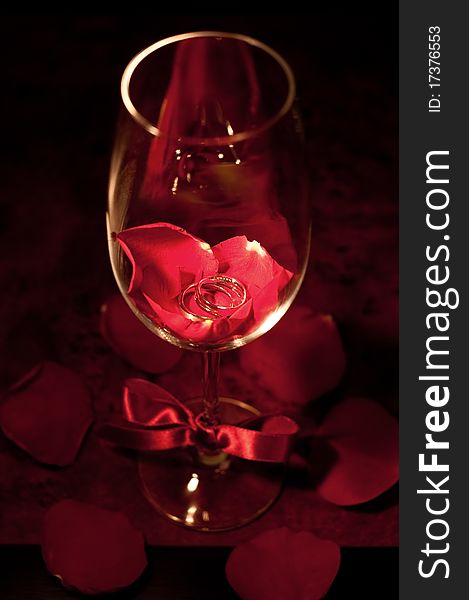 A pair of silver wedding ring in a red wine glass with fresh red rose petals underneath it. A pair of silver wedding ring in a red wine glass with fresh red rose petals underneath it.
