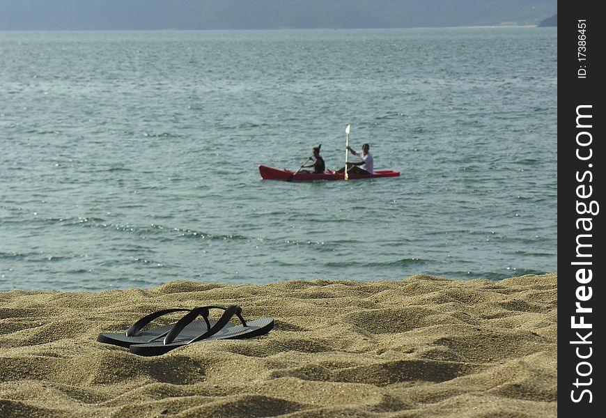 Summer flip-flop on the sand with the ocean at the background and two people on a kaiak. Summer flip-flop on the sand with the ocean at the background and two people on a kaiak