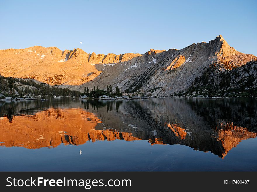 Moon rising over a mountain ridge glowing orange from the setting sun, reflected in a calm alpine lake. Moon rising over a mountain ridge glowing orange from the setting sun, reflected in a calm alpine lake.