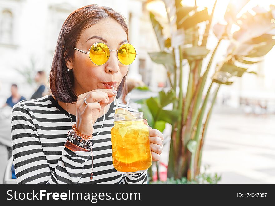 Happy female hipster student drinks a cool lemonade through a straw in a summer outdoor cafe