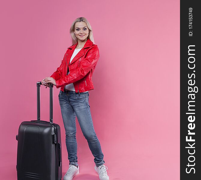 Young female tourist in a red jacket and with Luggage stands on a pink background. Tourist girl ready to fly, copy space. Young female tourist in a red jacket and with Luggage stands on a pink background. Tourist girl ready to fly, copy space