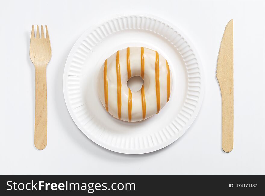 Glazed donut on a paper plate and wooden cutlery on a white background, top view. Glazed donut on a paper plate and wooden cutlery on a white background, top view