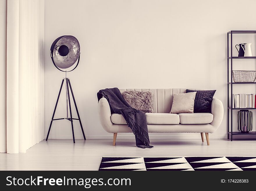 Industrial black lamp next to beige sofa with blanket and pillows and shelf with books, copy space on empty white wall