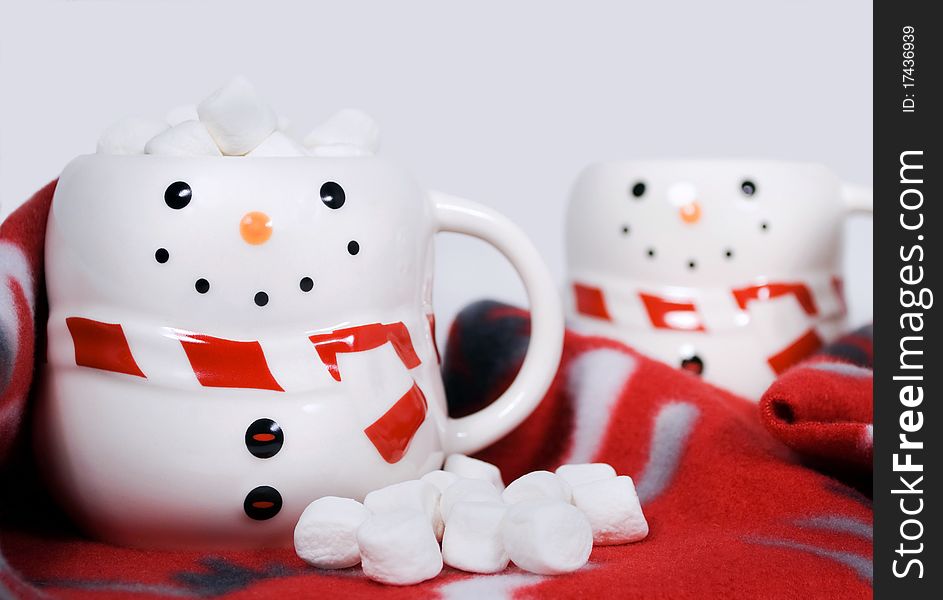 Two cute snowman mugs with hot chocolate and marshmallows on a cozy red winter scarf, focus on foreground snowman