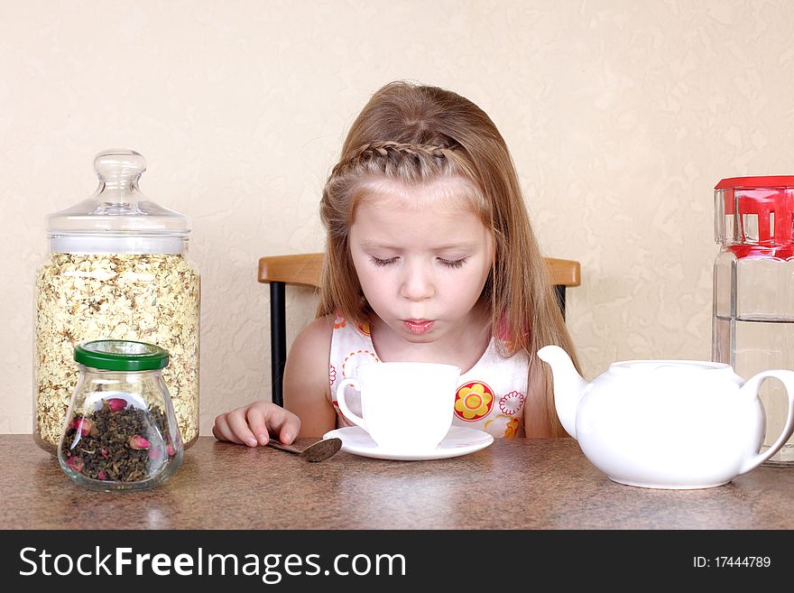 Little girl blowing to cup of hot drink, at table with lime, tea, jasmin petals and teapot against yellow wall in home