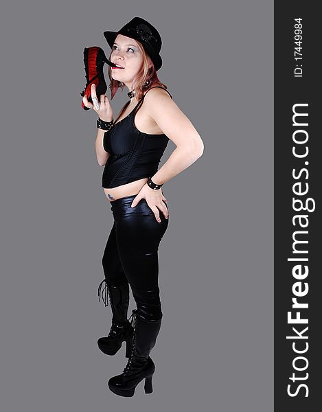 A young punk woman in black leather pants, black corset and boots and a hat standing with her back, for gray background. A young punk woman in black leather pants, black corset and boots and a hat standing with her back, for gray background.