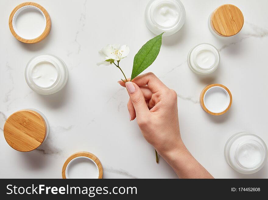 Cropped view of woman hand with jasmine flowers over jars with cosmetic cream on white surface