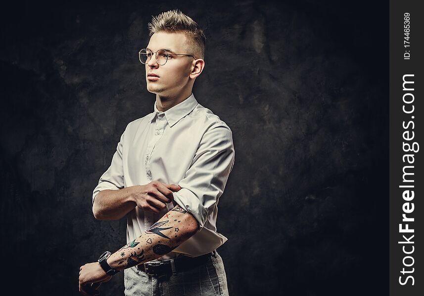 Young handsome student guy in a white shirt, with tattoos on his hand, glasses and watches, tucks up his sleeves in a dark studio isolated on grey background, looking confident and thinking. Young handsome student guy in a white shirt, with tattoos on his hand, glasses and watches, tucks up his sleeves in a dark studio isolated on grey background, looking confident and thinking