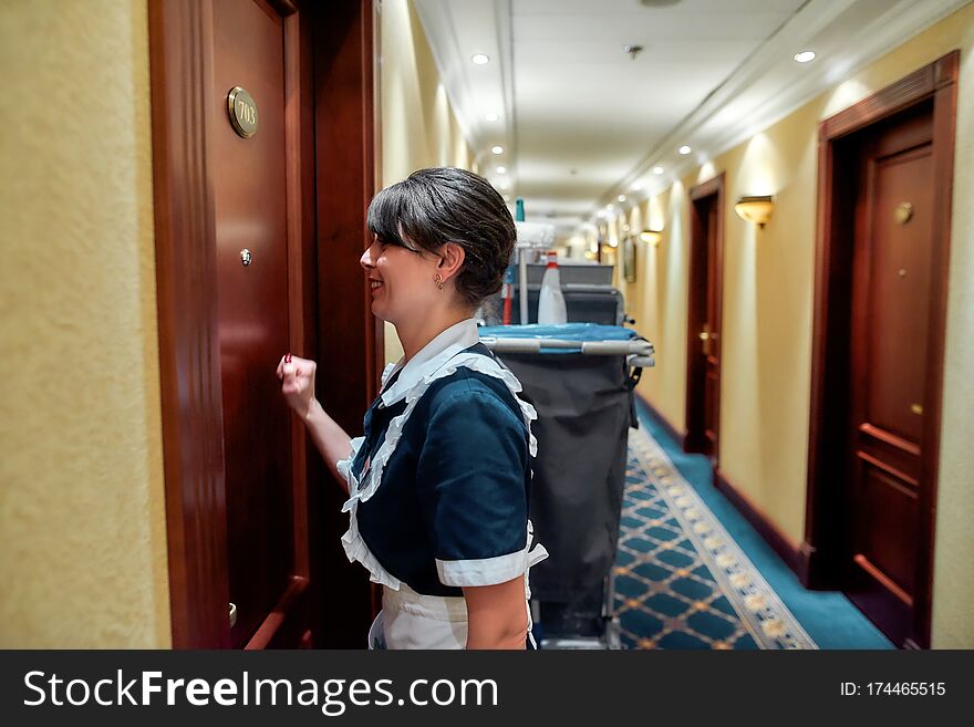 Smiling hotel maid in uniform knocking on the door for room service while standing with the chambermaid trolley in the hall. Horizontal shot. Side view. Smiling hotel maid in uniform knocking on the door for room service while standing with the chambermaid trolley in the hall. Horizontal shot. Side view