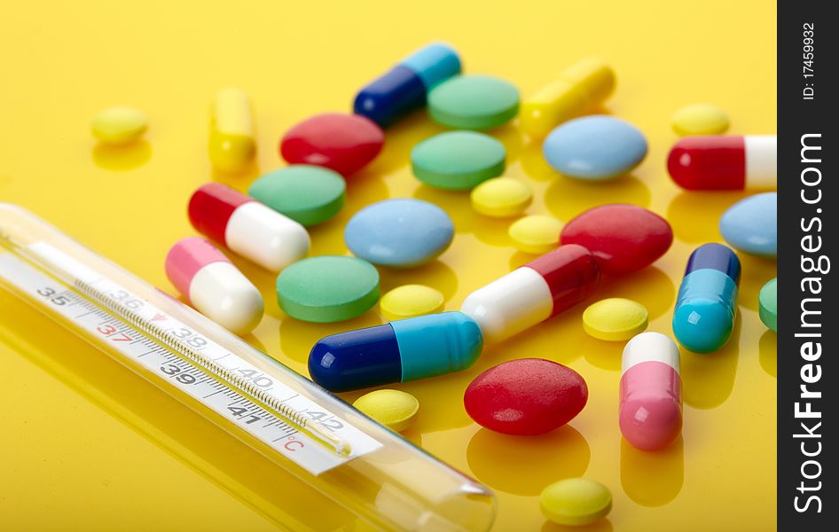 Colorful pills and thermometer over yellow background