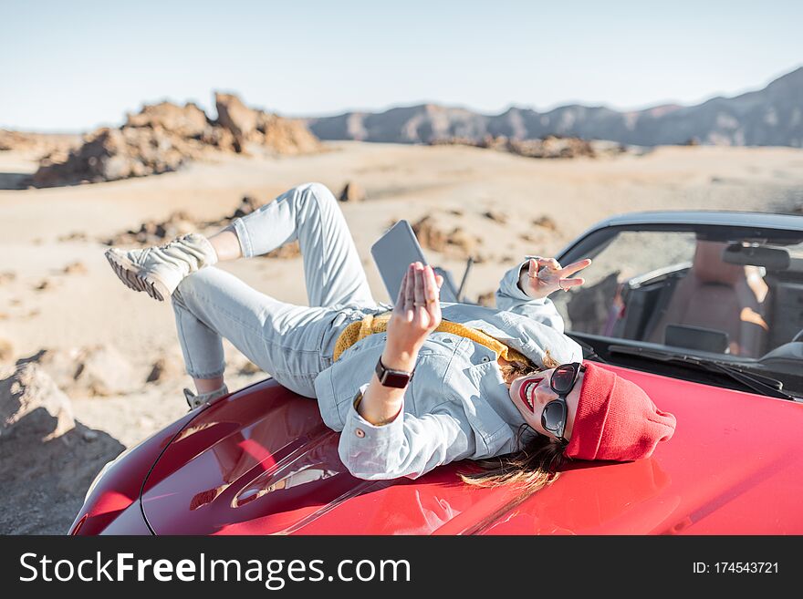 Lifestyle portrait of a young woman enjoying road trip on the desert valley, lying on the car hood and photographing on phone. Lifestyle portrait of a young woman enjoying road trip on the desert valley, lying on the car hood and photographing on phone