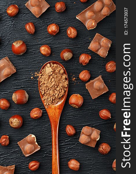 Chocolate with hazelnuts and a wooden spoon with cocoa on a dark background, surrounded by nuts in the shell and peeled. Still-life of Confectionery products. Top view, close-up