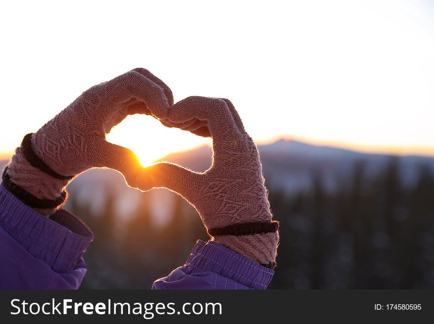 Woman making heart with her hands in mountains at sunset, closeup. Winter vacation