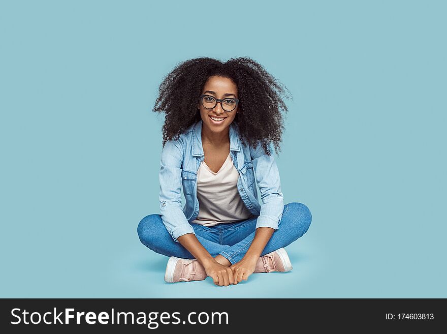 Young african girl wearing eyeglasses sitting on floor  on gray background leaning forward looking camera smiling joyful. Young african girl wearing eyeglasses sitting on floor  on gray background leaning forward looking camera smiling joyful