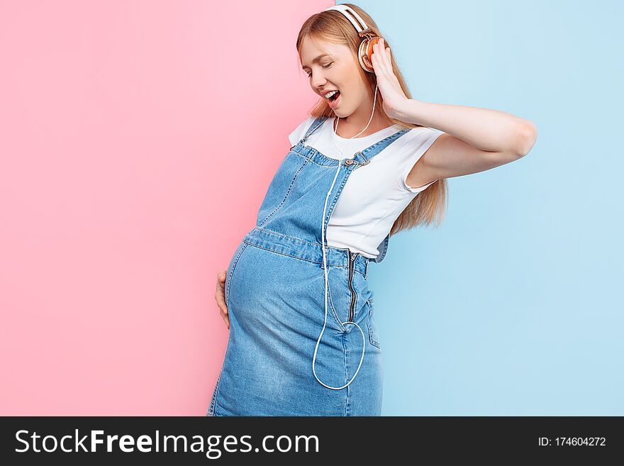 Happy cute beautiful young cheerful pregnant woman enjoys listening to music with headphones while standing on an  pink and blue background. Happy cute beautiful young cheerful pregnant woman enjoys listening to music with headphones while standing on an  pink and blue background