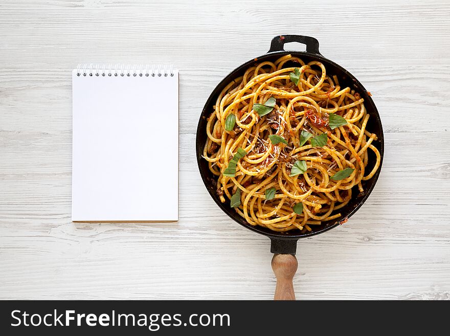 Homemade Bucatini all Amatriciana Pasta in a cast iron pan, blank notepad on a white wooden background, top view. Flat lay