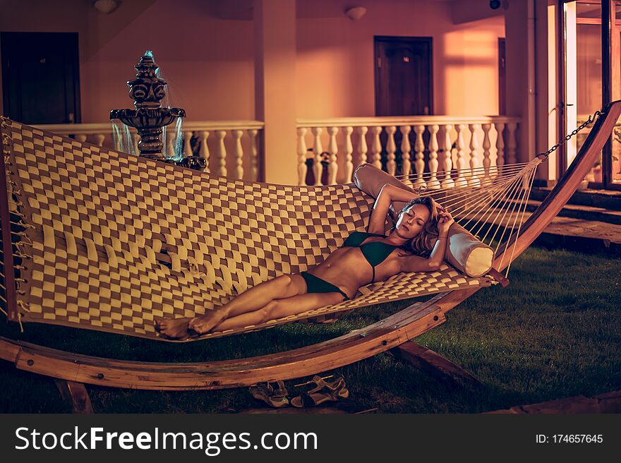 Slender beautiful young woman in a swimsuit is lying on a wicker hammock in the night yard of the garden of her house in summer. Slender beautiful young woman in a swimsuit is lying on a wicker hammock in the night yard of the garden of her house in summer