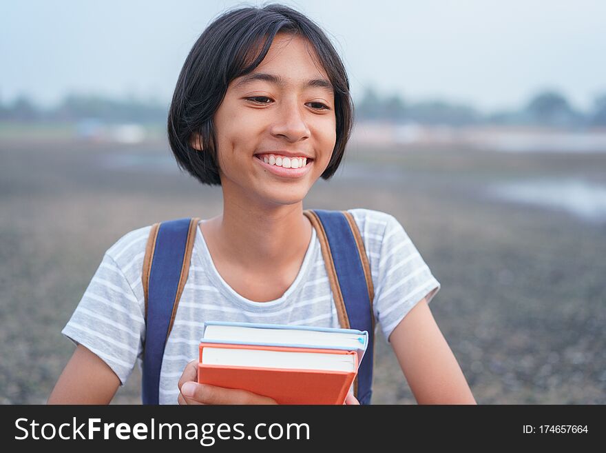 Happy Asian Girl Smile On Face And Laugh While Standing Amid Nature In The Morning, Asia Child Hold Book And Backpack On Blurred