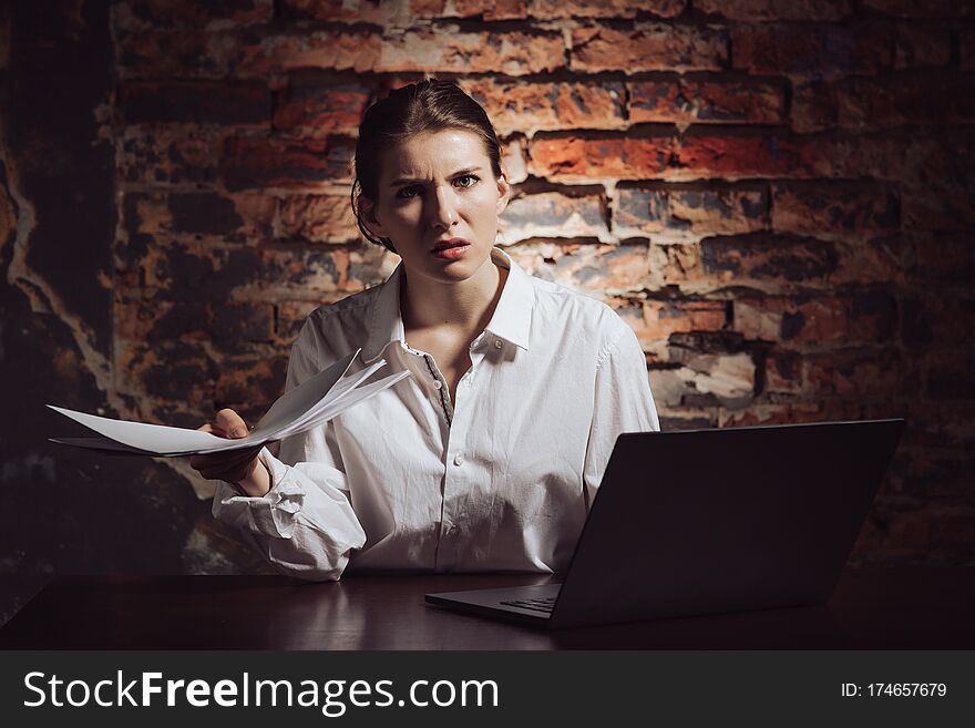 Displeased confused young female entrepreneur holding documents and looking inquiringly at camera while sitting at table with laptop against brick wall. Displeased confused young female entrepreneur holding documents and looking inquiringly at camera while sitting at table with laptop against brick wall