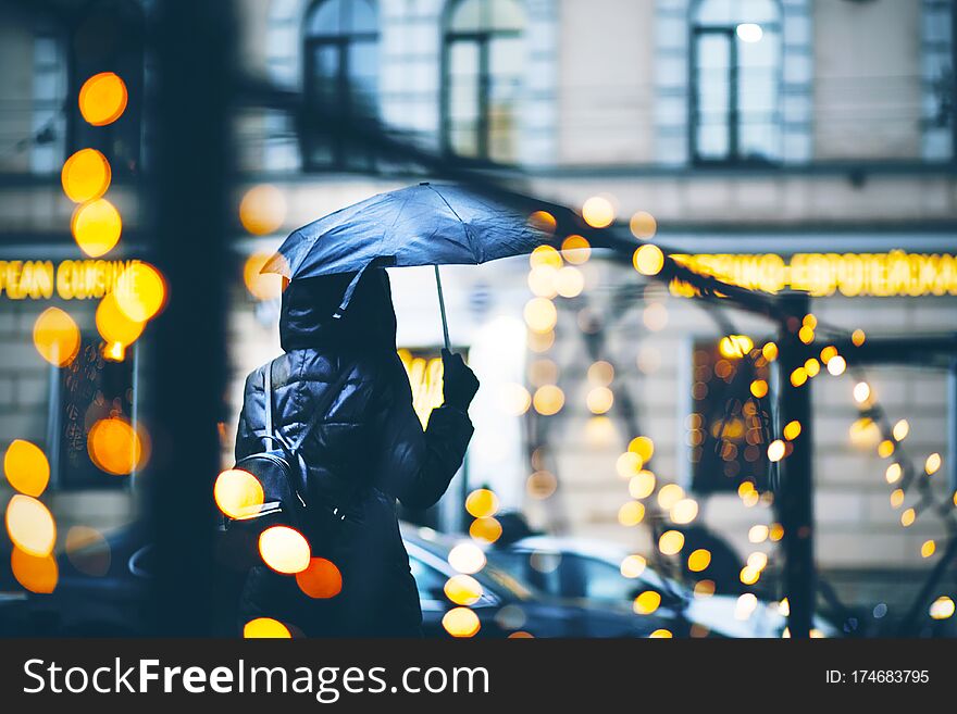 A girl in a black windbreaker with a wet umbrella walks along a city street in the evening, illuminated by lights that decorate the black fence. A girl in a black windbreaker with a wet umbrella walks along a city street in the evening, illuminated by lights that decorate the black fence