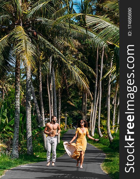 Beautiful Couple Running Among Palm Trees In Bali, Indonesia. Honeymoon On The Islands. A Happy Couple In Love Runs Along The Road