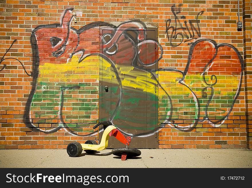 A broken child's tricycle is abandoned near an old storage warehouse that has been vandalized with graffiti. A broken child's tricycle is abandoned near an old storage warehouse that has been vandalized with graffiti.