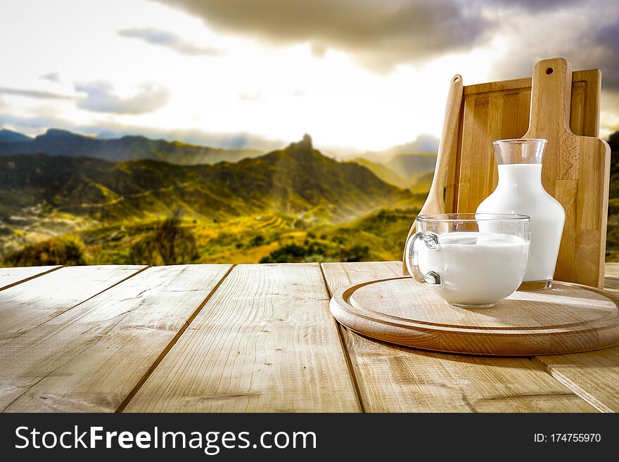 Wooden table top background with space for advertising product or decoration. White sky and mountains view in distance. Fresh milk in a glass and wooden cutting board on table top with mountains background and spring time. Wooden table top background with space for advertising product or decoration. White sky and mountains view in distance. Fresh milk in a glass and wooden cutting board on table top with mountains background and spring time.